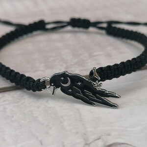 May include: A black braided bracelet with a silver charm featuring a stylized raven with a crescent moon and stars.