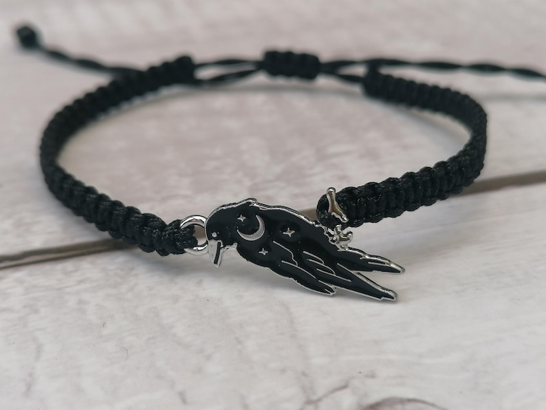 May include: A black braided bracelet with a silver charm featuring a stylized raven with a crescent moon and stars.