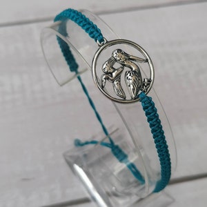 May include: Teal braided bracelet with a silver charm featuring two pelicans in a circle.