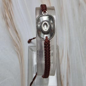 May include: A brown macrame bracelet featuring a silver-toned cowboy hat charm. The hat charm has a rope-like border and an oval center. The bracelet is displayed on a clear stand against a neutral background.