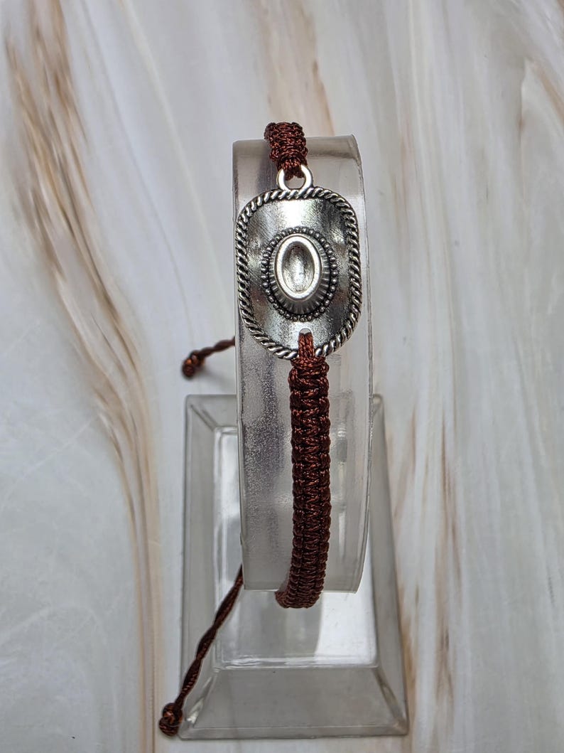 May include: A brown macrame bracelet featuring a silver-toned cowboy hat charm. The hat charm has a rope-like border and an oval center. The bracelet is displayed on a clear stand against a neutral background.