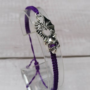 May include: Purple friendship bracelet with a silver charm of a dragon with wings.