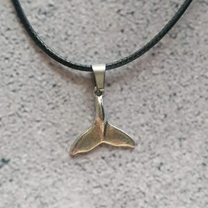 Könnte beinhalten: Ein silberfarbener Walflossen-Anhänger hängt an einer schwarzen Kordelkette. Der Anhänger ist ein detailliertes, dreidimensionales Design einer Walflosse. Die Halskette wird vor einem strukturierten, grauen Hintergrund präsentiert.