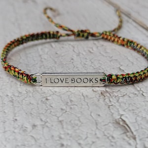 Puede incluir: Una pulsera trenzada colorida con un dije plateado que dice "ME ENCANTAN LOS LIBROS".