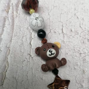 Puede incluir: Un charm de oso marrón con una oreja amarilla y un charm de estrella marrón con detalles dorados. Los charms están unidos a un cordón marrón con cuentas transparentes y negras.
