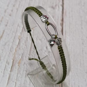 May include: Olive green macrame bracelet with silver acorn and leaf charms. The bracelet has an adjustable sliding knot closure. The charms feature leaf and acorn designs. The bracelet is displayed on a clear plastic stand.