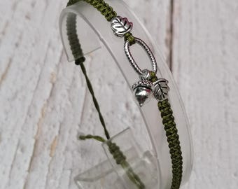 May include: Olive green macrame bracelet with silver-toned acorn and leaf charms. The bracelet features a sliding knot closure for adjustable sizing. The charms are detailed with leaf and acorn designs. The bracelet is displayed on a clear plastic stand.