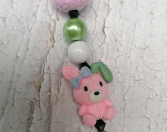 Könnte beinhalten: Ein rosa Hasen-Anhänger mit einer blauen Schleife und grünen Ohren, aufgereiht auf einer schwarzen Schnur mit rosa, grünen und weißen Perlen.