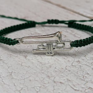 Könnte beinhalten: Ein Armband aus dunkelgrünem geflochtenem Kordelmaterial mit einem silberfarbenen Trompetenanhänger aus Metall in der Mitte. Das Armband verfügt über verstellbare Schiebeknoten auf der Rückseite für eine sichere Passform und ein detailliertes Design.