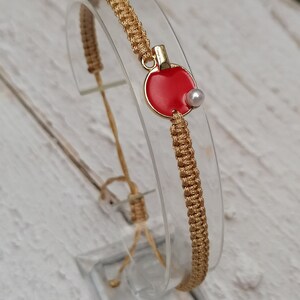 May include: A gold macrame bracelet with a red and gold ping pong paddle charm and a small white pearl. The bracelet is displayed on a clear stand against a white wooden background.
