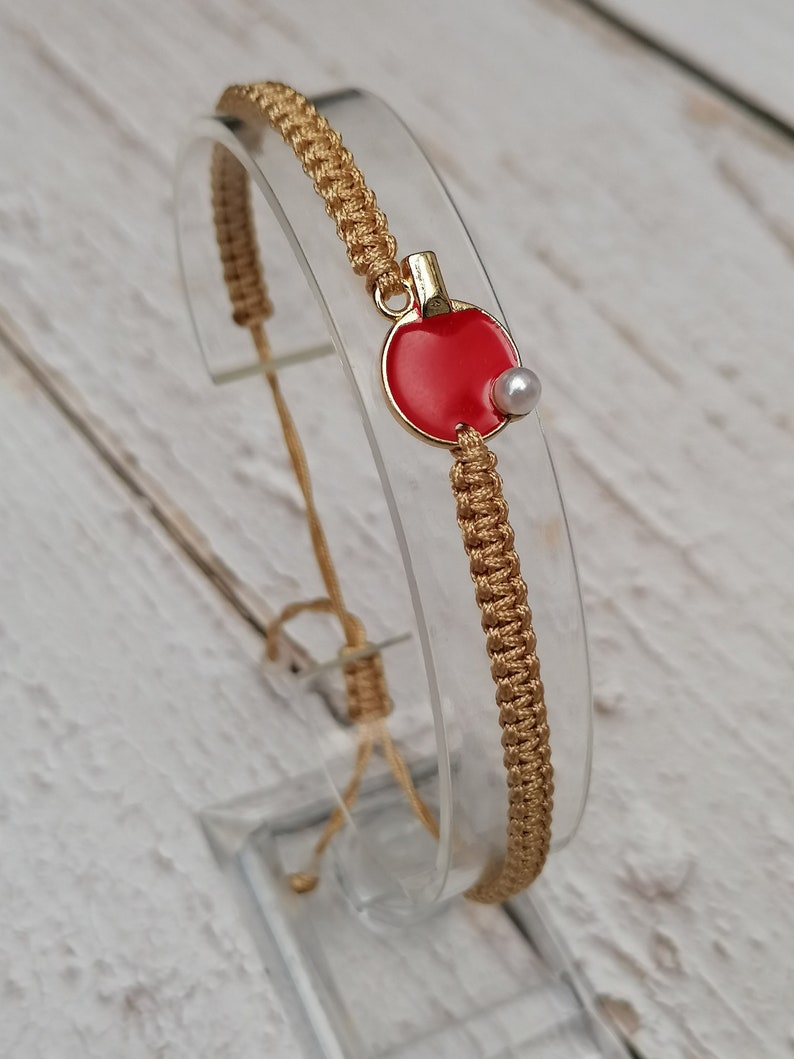 May include: A gold macrame bracelet with a red and gold ping pong paddle charm and a small white pearl. The bracelet is displayed on a clear stand against a white wooden background.