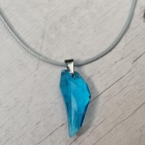 Puede incluir: Collar con colgante de cristal azul. El colgante tiene forma facetada e irregular, con un cierre de metal plateado. El cordón del collar es azul claro y el fondo es un veteado de madera clara.