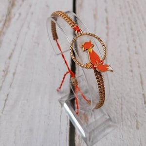 Könnte beinhalten: Ein geflochtenes Armband in Braun und Rot mit einem goldenen Anhänger, der eine rote Fuchs-Silhouette zeigt.