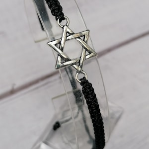 May include: A black cord bracelet featuring a silver-toned Star of David charm. The bracelet is constructed with a braided macrame-style knot pattern and includes a central metal pendant shaped as a six-pointed star with a textured finish.