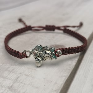 Può includere: Un bracciale in corda marrone scuro con un ciondolo in metallo color argento a forma di foglia di quercia con due piccole ghiande. Il bracciale presenta una chiusura a nodo scorsoio regolabile con due estremità libere.