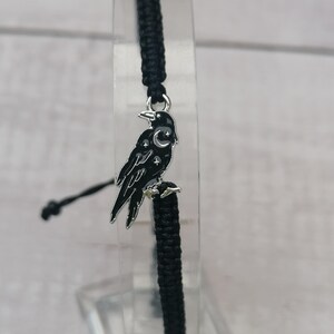May include: Black braided bracelet with a silver charm of a crow with a crescent moon and stars on its chest.