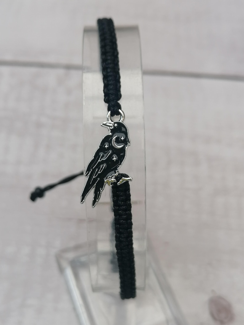 May include: Black braided bracelet with a silver charm of a crow with a crescent moon and stars on its chest.
