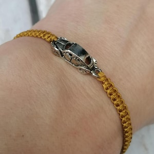 Puede incluir: Una pulsera de macram&eacute; dorada con un charm de coche plateado. El charm es una miniatura detallada de un coche cl&aacute;sico. La pulsera se lleva en la mu&ntilde;eca, mostrando el intrincado anudado del cord&oacute;n.