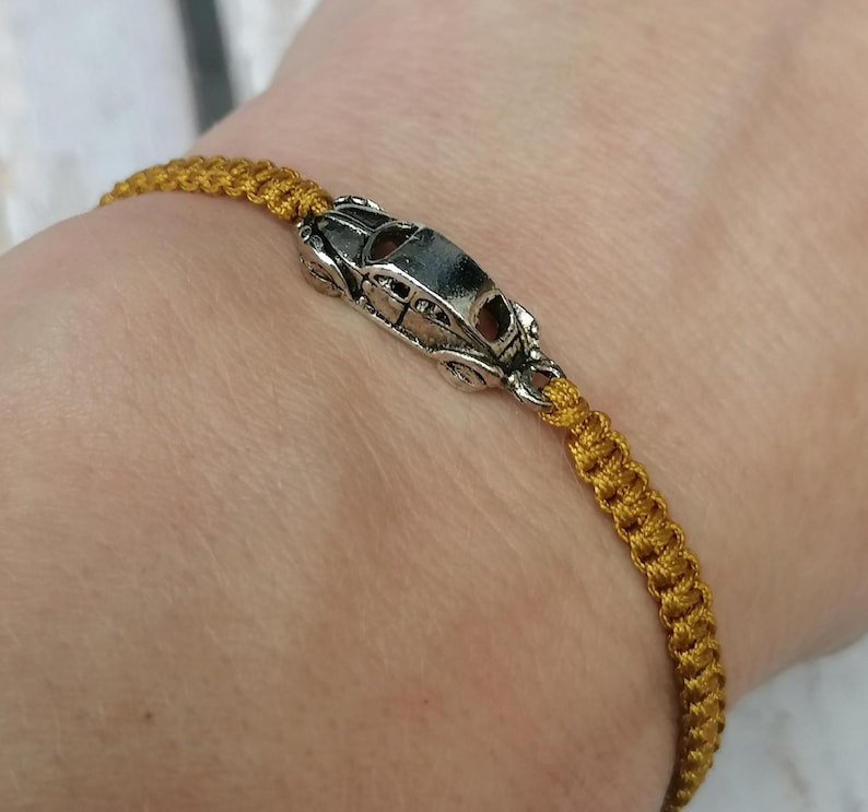 Puede incluir: Una pulsera de macram&eacute; dorada con un charm de coche plateado. El charm es una miniatura detallada de un coche cl&aacute;sico. La pulsera se lleva en la mu&ntilde;eca, mostrando el intrincado anudado del cord&oacute;n.