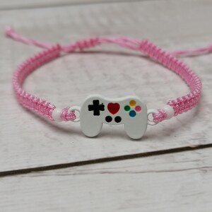 May include: A pink braided bracelet with a white plastic video game controller charm. The controller has a red heart and colourful buttons.