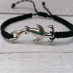 K&ouml;nnte beinhalten: Ein schwarzes geflochtenes Armband mit einem silbernen Hammerhai-Anh&auml;nger.