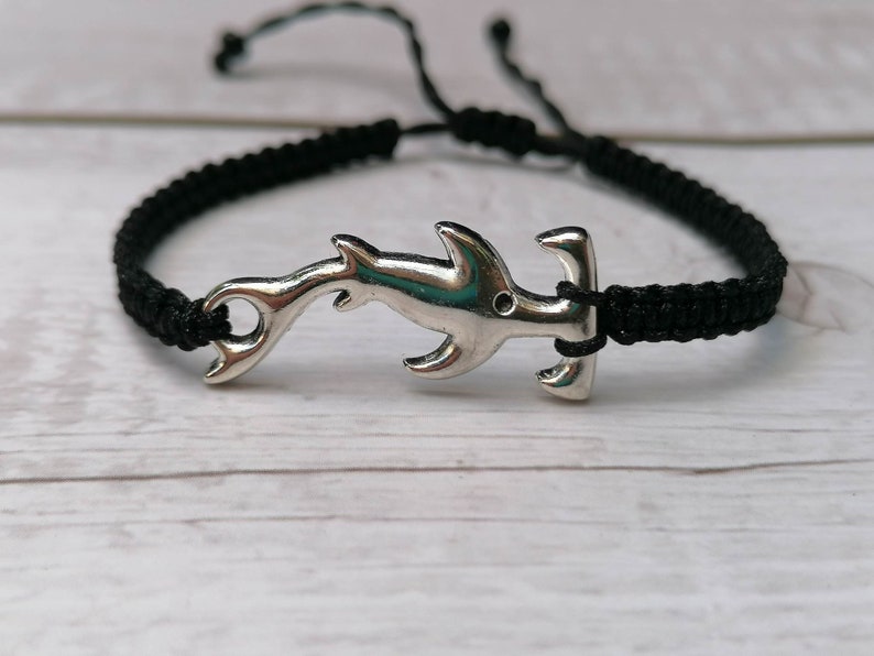 K&ouml;nnte beinhalten: Ein schwarzes geflochtenes Armband mit einem silbernen Hammerhai-Anh&auml;nger.