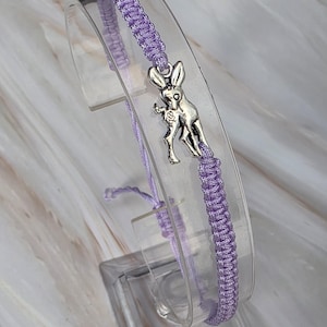 Könnte beinhalten: Ein lavendelfarbenes Kordelarmband mit einem silberfarbenen Metallanhänger in Form eines Esels. Das Armband ist mit einem geflochtenen Knotenmuster gefertigt und verfügt über verstellbare Kordelenden. Der Metallanhänger zeigt einen stehenden Esel im Profil.