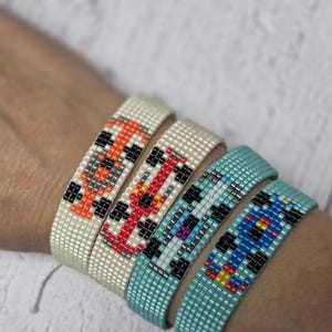 Könnte beinhalten: Vier Armbänder mit Perlen, jedes mit einem anderen Farbschema. Das erste Armband ist cremefarben mit orangefarbenen, schwarzen und grauen Perlen. Das zweite ist cremefarben mit roten, schwarzen und weißen Perlen. Das dritte ist türkis mit schwarzen, weißen und lila Perlen. Das vierte ist türkis mit blauen, gelben und schwarzen Perlen.