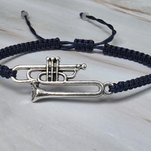 May include: A dark blue cord bracelet featuring a silver-toned metal trumpet charm. The bracelet has an adjustable woven band with two small beads at the ends of the pull strings, allowing for a customizable fit around the wrist.