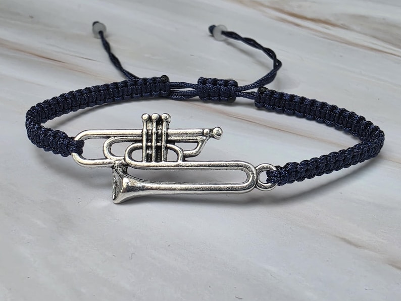 May include: A dark blue cord bracelet featuring a silver-toned metal trumpet charm. The bracelet has an adjustable woven band with two small beads at the ends of the pull strings, allowing for a customizable fit around the wrist.