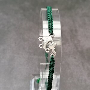 May include: A dark green woven cord bracelet featuring a silver-toned metal charm shaped like a pair of roller skates. The bracelet is constructed with a braided nylon cord and includes a central metallic skate pendant as the focal point.