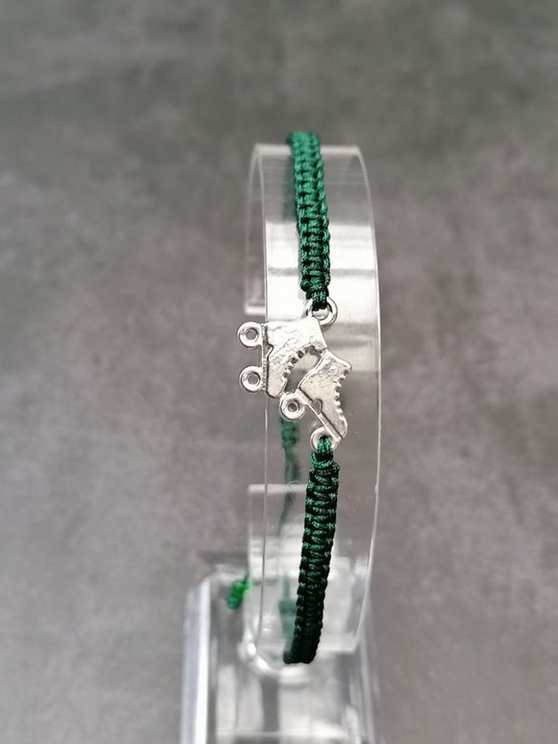 May include: A dark green woven cord bracelet featuring a silver-toned metal charm shaped like a pair of roller skates. The bracelet is constructed with a braided nylon cord and includes a central metallic skate pendant as the focal point.