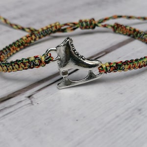 May include: A colourful braided bracelet featuring a silver ice skate charm. The bracelet is made of green, yellow, orange, and brown threads. The ice skate charm is detailed and has a classic design.