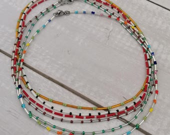 May include: Five colorful beaded necklaces with silver metal clasps. The necklaces are layered on a white wooden surface.