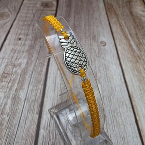 Puede incluir: Una pulsera de macramé amarilla con un dije de piña plateada. La pulsera se exhibe sobre un soporte de plástico transparente contra un fondo de madera. El dije de piña está detallado con una superficie texturizada.