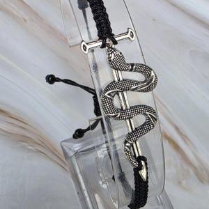 May include: A black cord bracelet featuring a silver-toned metal charm of a snake coiled around a sword. The bracelet has a braided texture and includes adjustable pull strings with small black knots at the ends to secure the fit on the wrist.