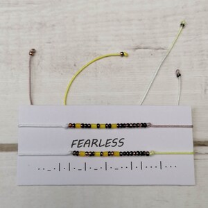 Puede incluir: Dos pulseras de cuentas en una tarjeta blanca con la palabra "FEARLESS" impresa en ella. Las pulseras están hechas con cuentas negras, amarillas y de cobre en hilo blanco, rosa claro y amarillo claro.