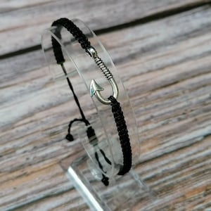 Puede incluir: Un brazalete trenzado negro con un dije de anzuelo de pesca plateado. El brazalete se muestra en un soporte transparente.