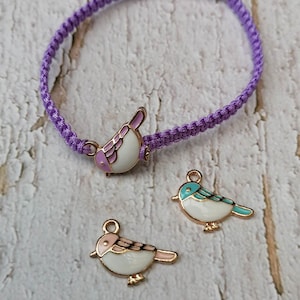 May include: A purple braided bracelet with a white bird charm with pink and purple accents. Two additional charms are shown, one with a blue bird and one with a pink bird.