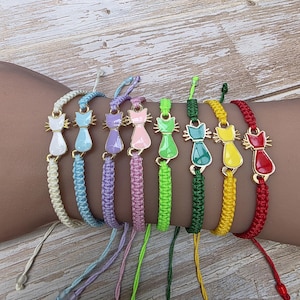 May include: A selection of cat charm bracelets in various colours. Each bracelet has a gold-coloured cat charm with enamel details in colours such as white, blue, purple, pink, green, yellow, and red. The bracelets are made with braided cords in matching colours.