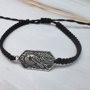 May include: A black cord bracelet featuring an adjustable braided band and a central silver-toned hexagonal charm. The charm displays a detailed embossed design of a bird perched on a branch against a circular moon background with intricate textures.