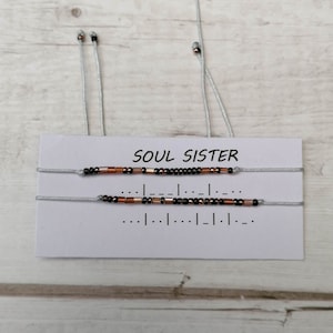 Könnte beinhalten: Zwei Armbänder mit Perlen auf silbernem Band, mit einer Karte mit der Aufschrift "SOUL SISTER". Die Armbänder haben schwarze und kupferfarbene Perlen. Die Karte zeigt auch eine Morsecode-Nachricht.