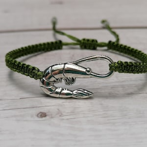 May include: A green braided cord bracelet with a silver shrimp charm. The bracelet has a lobster clasp closure.