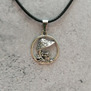 May include: Silver-toned pendant necklace featuring a mushroom and owl design within a circular frame. The pendant hangs from a black cord. The mushroom has a dotted cap. The owl is perched below the mushroom.