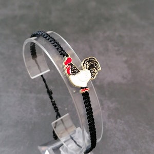 May include: Black braided bracelet featuring a rooster charm. The charm is white with black spots, a red comb, and a black and white tail. The bracelet is displayed on a clear stand against a gray background.