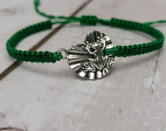 Könnte beinhalten: Ein grünes Armband aus geflochtener Kordel mit einem zentralen silberfarbenen Metallanhänger in Form eines Frosches auf einem Seerosenblatt. Das Armband verfügt über verstellbare Schiebeknoten und ein geflochtenes Design mit einer detaillierten Figur.