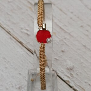 May include: A gold-colored macrame bracelet with a red circular charm. The charm has a gold border and a small white pearl. The bracelet is displayed on a clear stand against a light wood background.