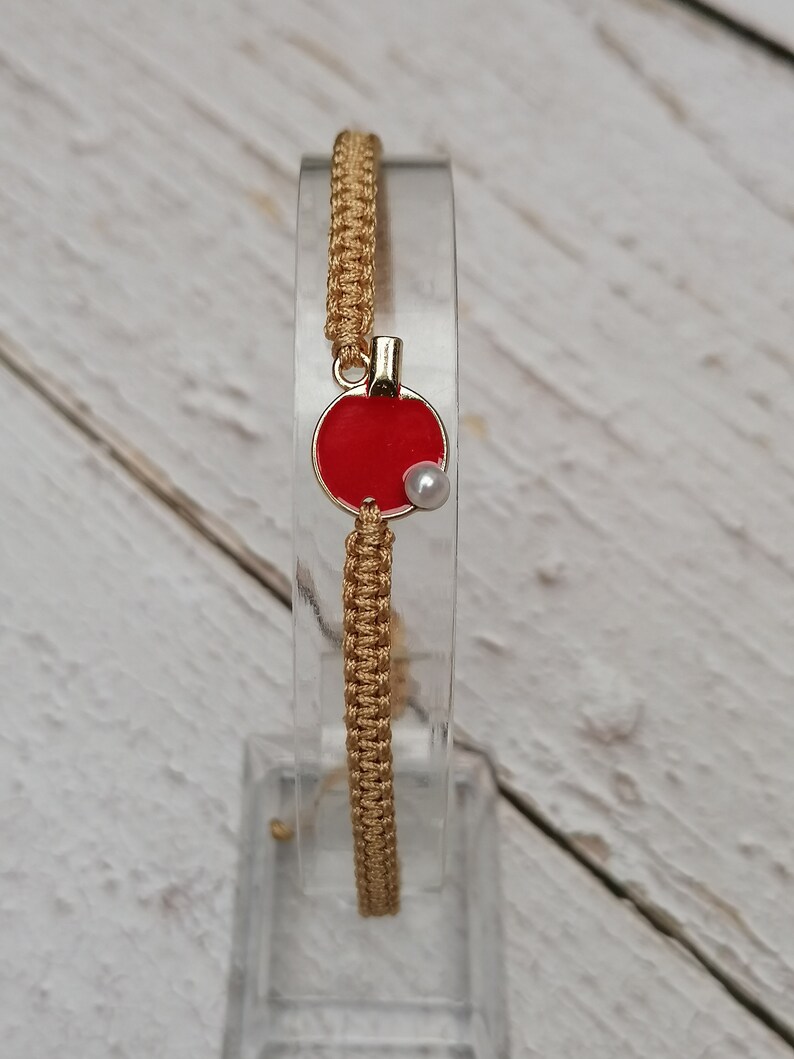 May include: A gold-colored macrame bracelet with a red circular charm. The charm has a gold border and a small white pearl. The bracelet is displayed on a clear stand against a light wood background.