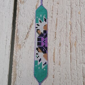 May include: A beaded bookmark with a teal background and a pattern of white, black, tan, and purple squares. The bookmark has a braided purple cord attached at the top.