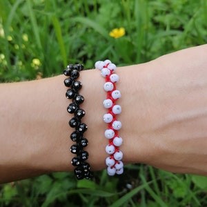 May include: Two beaded bracelets, one black and one white, with red string accents. The bracelets are worn on a person's wrist.